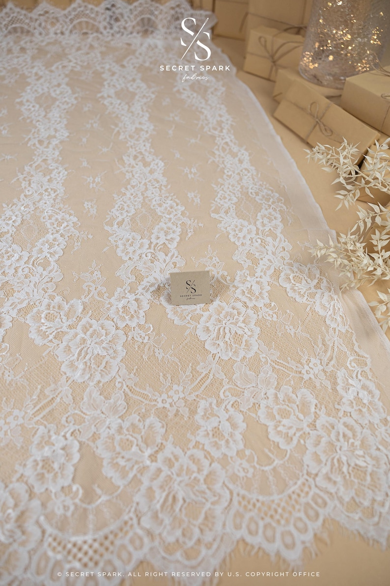 May include: White floral lace fabric with a delicate pattern. The lace is laid out on a beige surface. The image includes the text "SECRET SPARK fabrics" and "SECRET SPARK. ALL RIGHT RESERVED BY U.S. COPYRIGHT OFFICE."