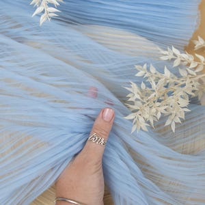 May include: Light blue tulle fabric with a pleated texture. The fabric is being held by a hand wearing a silver ring that says "love".