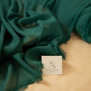 May include: A close-up of a deep green fabric with a frayed edge. The fabric is folded and draped on a light brown surface. A small white card with the text "SECRET SPARK fabrics" is visible in the foreground.