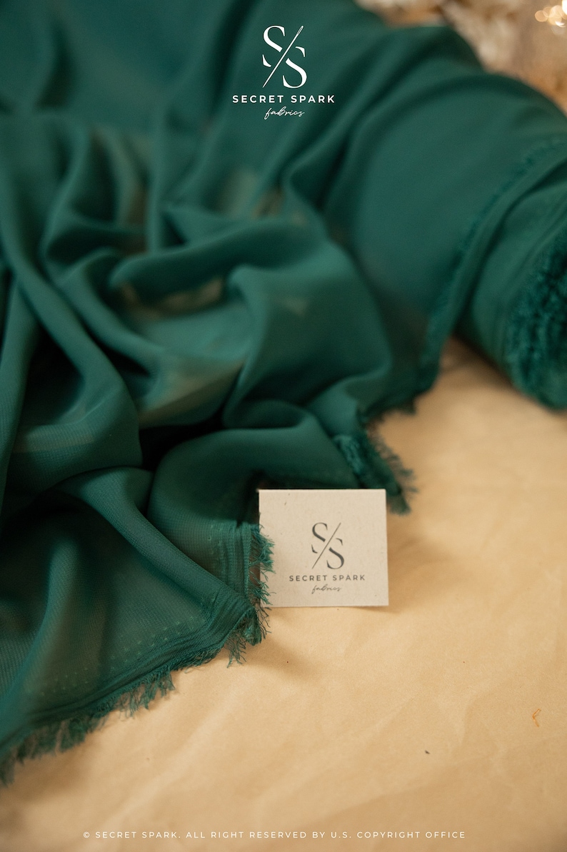 May include: A close-up of a deep green fabric with a frayed edge. The fabric is folded and draped on a light brown surface. A small white card with the text "SECRET SPARK fabrics" is visible in the foreground.