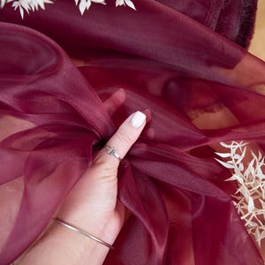 May include: A close-up of a hand holding a burgundy sheer fabric. The fabric is translucent and has a slight sheen. The hand is wearing a silver ring.
