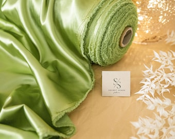 May include: A roll of light green satin fabric with a fringed edge. The fabric is rolled up and tied with a white ribbon. The fabric is sitting on a brown surface with a white tag that says "Secret Spark Fabrics".