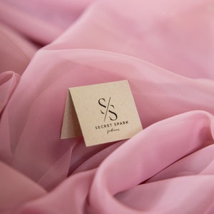May include: A close-up of a small, brown, rectangular tag with the text "S/S SECRET SPARK fabrics" printed in black. The tag is lying on a pink fabric.