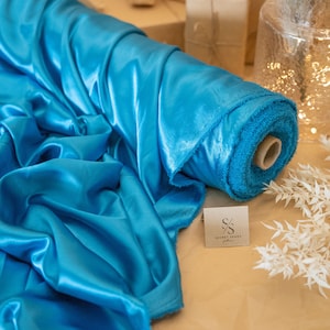 May include: A roll of vibrant turquoise satin fabric, with a soft, lustrous sheen. The fabric is displayed with a small card that reads "SECRET SPARK fabrics". The image showcases the fabric's texture and color, ideal for apparel or decor.