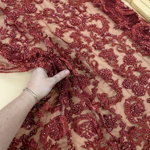 May include: Burgundy lace fabric with floral and beaded embellishments. The delicate design features intricate patterns and textures, perfect for bridal wear or special occasion garments. The fabric is semi-transparent.