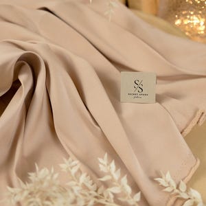 May include: A close-up of a light beige fabric with a soft, smooth texture. The fabric is folded and draped, creating a luxurious and elegant look. The fabric is labelled with the text "SECRET SPARK fabrics".