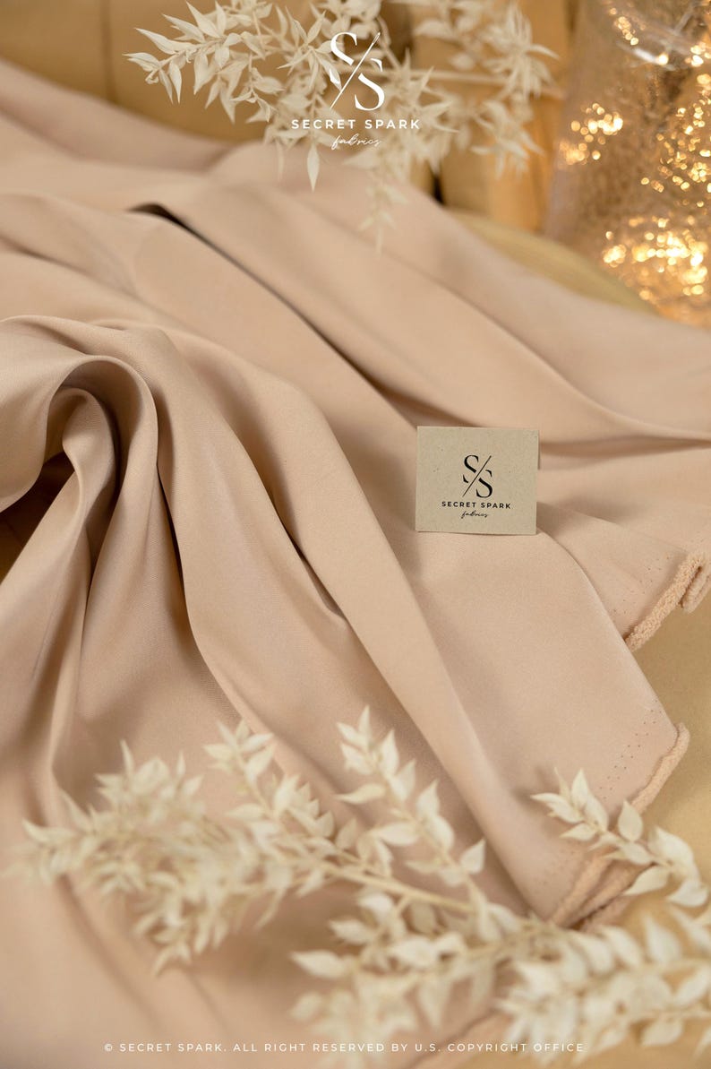 May include: A close-up of a light beige fabric with a soft, smooth texture. The fabric is folded and draped, creating a luxurious and elegant look. The fabric is labelled with the text "SECRET SPARK fabrics".