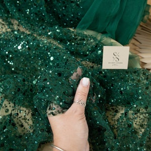 May include: Emerald green fabric with sequins and beads. The fabric is sheer and has a delicate texture. The fabric is folded in half and a hand is holding the fabric. The fabric is labeled "Secret Spark Fabrics".