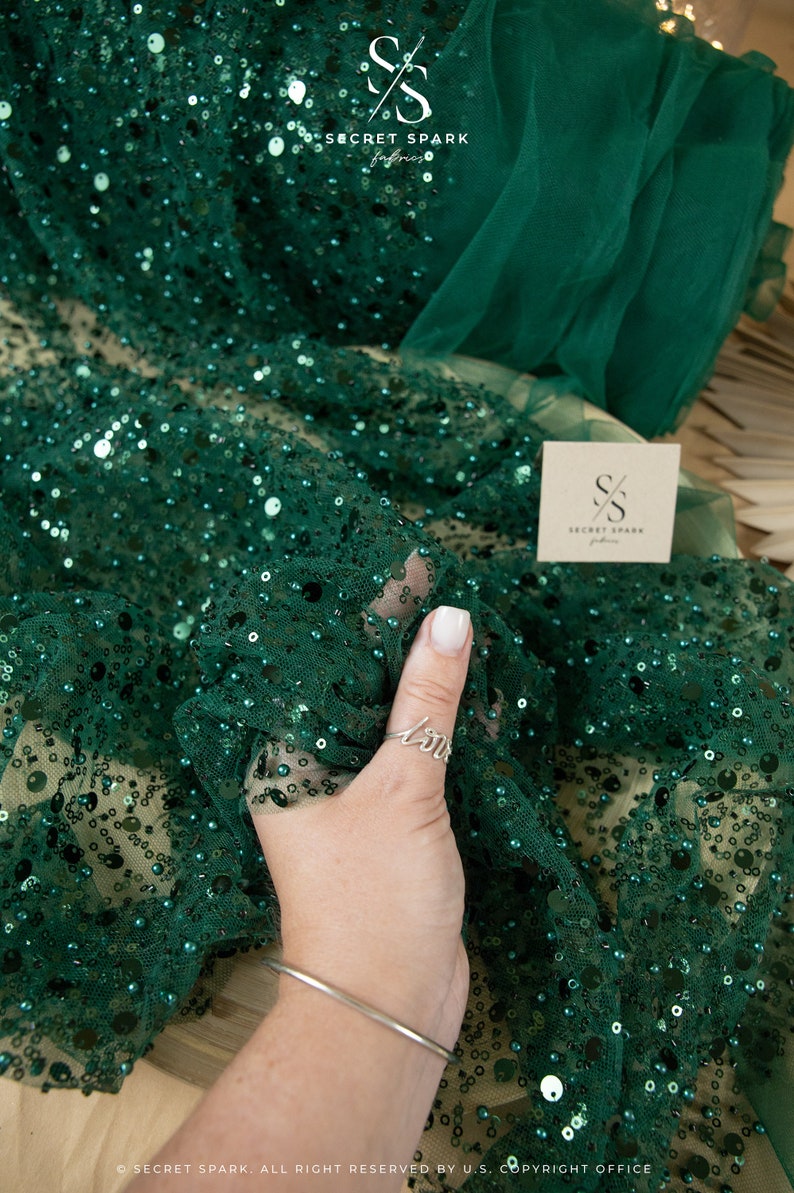 May include: Emerald green fabric with sequins and beads. The fabric is sheer and has a delicate texture. The fabric is folded in half and a hand is holding the fabric. The fabric is labeled "Secret Spark Fabrics".