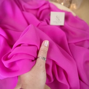 Puede incluir: Primer plano de una tela de color rosa brillante que se sostiene con una mano. La tela es transparente y tiene un ligero brillo. El texto "SECRET SPARK fabrics" es visible en la esquina superior izquierda de la imagen.
