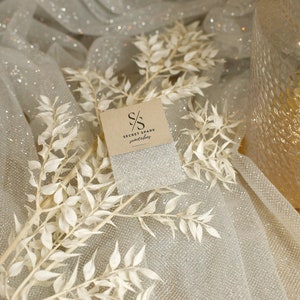 May include: A close-up of fabric swatches with the text "SECRET SPARK fabrics". The fabric is a shimmering silver with a glittery texture. White floral branches and a decorative glass vase are also in the image.