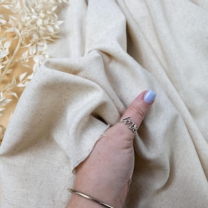 May include: A piece of off-white, textured fabric with visible flecks, featuring a silver-toned ring with the word love in cursive script and a simple, thin, smooth silver-toned metal bangle bracelet worn on the wrist.