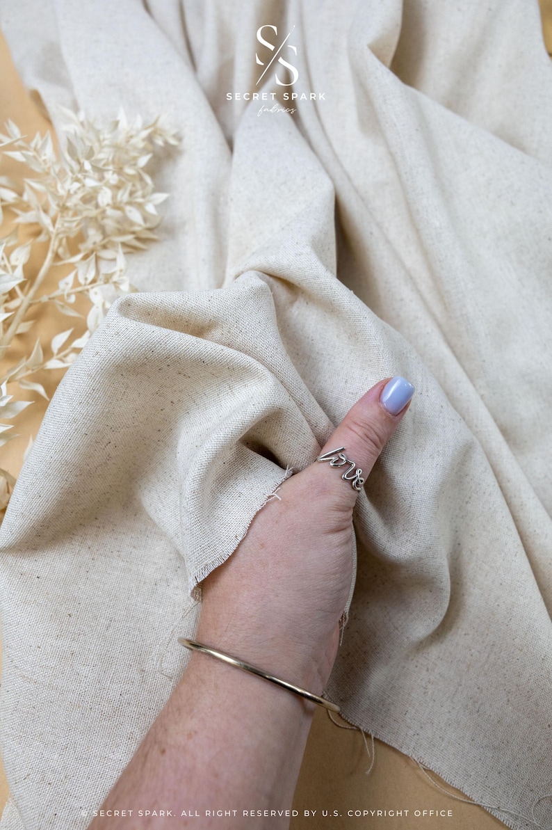 May include: A piece of off-white, textured fabric with visible flecks, featuring a silver-toned ring with the word love in cursive script and a simple, thin, smooth silver-toned metal bangle bracelet worn on the wrist.