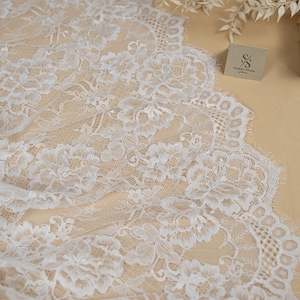 May include: White floral lace fabric with a delicate pattern. The lace is displayed on a beige surface, with the "SECRET SPARK" logo visible. The fabric's intricate design and texture are highlighted in this image.