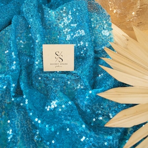 May include: Turquoise blue sequin fabric with a small white card that says "SECRET SPARK fabrics" in black text.