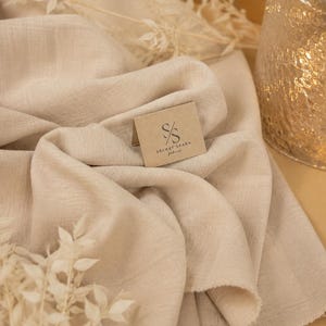 May include: A piece of beige fabric with a textured, woven appearance. A small, folded tan card rests on the fabric, featuring the logo and text 'SS SECRET SPARK fabrics'. The fabric has soft, draped folds and a visible, slightly frayed edge.