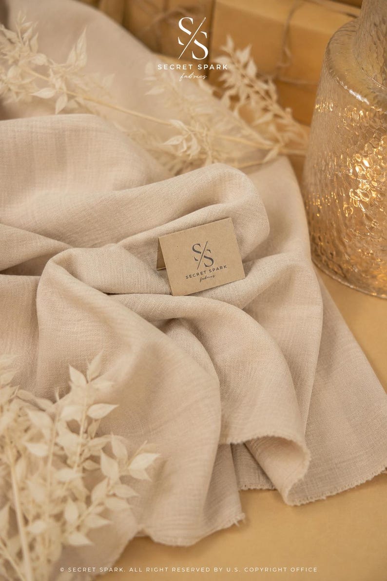 May include: A piece of beige fabric with a textured, woven appearance. A small, folded tan card rests on the fabric, featuring the logo and text 'SS SECRET SPARK fabrics'. The fabric has soft, draped folds and a visible, slightly frayed edge.