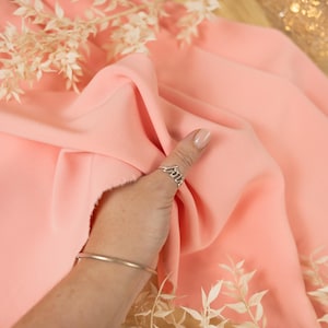 May include: A close-up of a peach-colored fabric being held by a hand. The fabric is smooth and soft, and the hand is wearing a silver ring. The fabric is on a table with a light brown surface. The text "SECRET SPARK fabrics" is visible in the top center of the image.