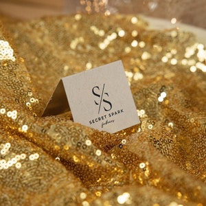 May include: A close-up of a small, brown card with the text "SECRET SPARK fabrics" and a stylized "SS" logo. The card is lying on a gold sequined fabric.