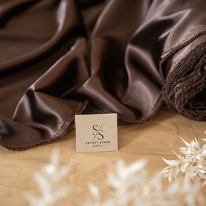 May include: A close-up of a brown satin fabric with a small brown paper tag that says "SECRET SPARK fabrics". The fabric is folded and draped over a brown surface.
