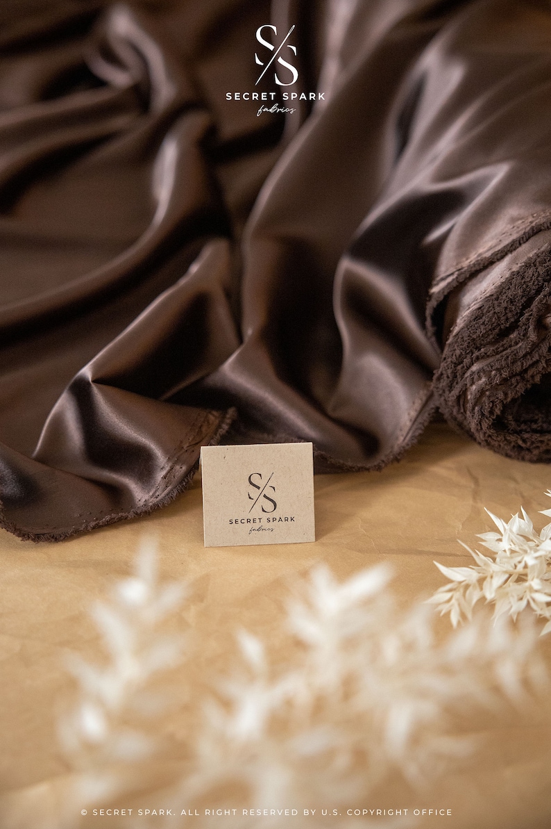May include: A close-up of a brown satin fabric with a small brown paper tag that says "SECRET SPARK fabrics". The fabric is folded and draped over a brown surface.