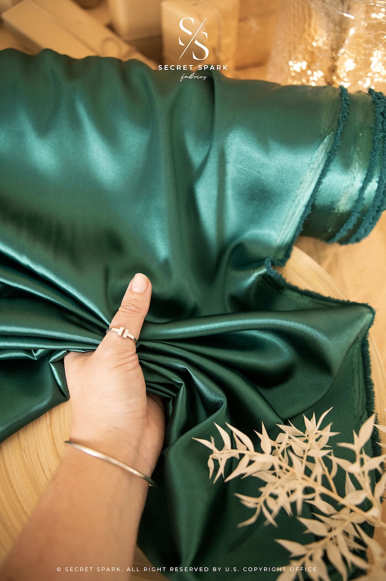 Puede incluir: Un primer plano de una tela de sat&eacute;n verde oscuro con una textura suave y sedosa. La tela est&aacute; doblada y drapeada sobre una superficie de madera. La tela es de un color rico y precioso.