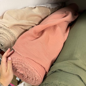 May include: Multiple rolls of fabric in various colors including white, beige, dusty rose, and olive green. The fabric has a visible crinkled texture and raw, frayed edges on the ends of the rolls, which are stacked horizontally on a shelf.