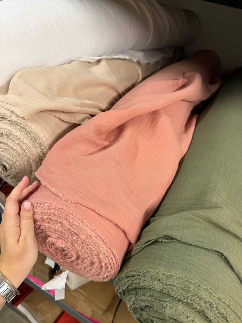 May include: Multiple rolls of fabric in various colors including white, beige, dusty rose, and olive green. The fabric has a visible crinkled texture and raw, frayed edges on the ends of the rolls, which are stacked horizontally on a shelf.