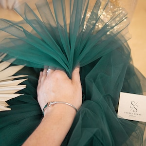 May include: A close-up of a green tulle fabric, folded in a fan shape. The fabric is soft and delicate, with a subtle sheen. The fabric is labeled "SECRET SPARK fabrics".