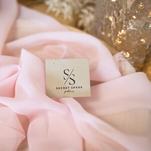 K&ouml;nnte beinhalten: Nahaufnahme eines hellrosa Stoffes mit einem kleinen, braunen, quadratischen Etikett, auf dem in schwarzer Schrift "SECRET SPARK fabrics" steht. Das Etikett hat ein stilisiertes "SS"-Logo.