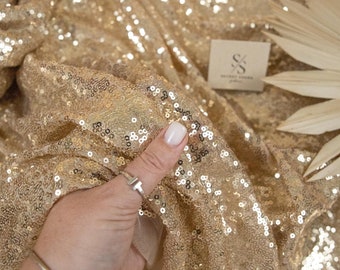 May include: Gold sequin fabric with a shimmering effect. The fabric is draped, showcasing its texture and light reflection. The image includes the "SECRET SPARK fabrics" logo.