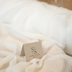 K&ouml;nnte beinhalten: Ein wei&szlig;er Stoffhintergrund mit einer kleinen, braunen Karte, auf der das Logo "SECRET SPARK fabrics" mit den sich &uuml;berlappenden Buchstaben "S" und einer diagonalen Linie durch sie hindurch abgebildet ist.