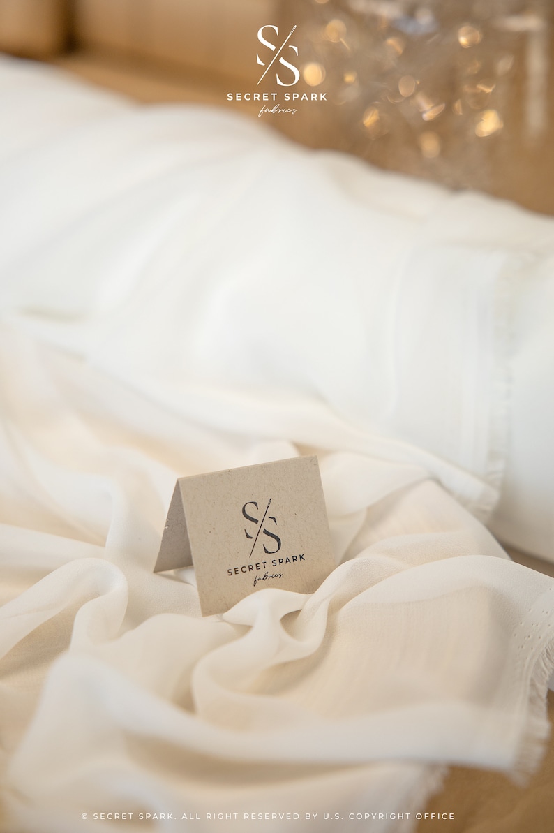 K&ouml;nnte beinhalten: Ein wei&szlig;er Stoffhintergrund mit einer kleinen, braunen Karte, auf der das Logo "SECRET SPARK fabrics" mit den sich &uuml;berlappenden Buchstaben "S" und einer diagonalen Linie durch sie hindurch abgebildet ist.