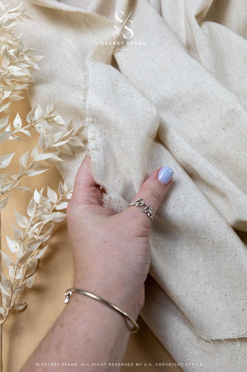 May include: A piece of off-white, textured fabric with visible flecks and a raw, frayed edge. The fabric is draped in soft folds. A logo at the top reads SECRET SPARK fabrics, and text at the bottom reads SECRET SPARK. ALL RIGHT RESERVED BY U.S. COPYRIGHT OFFICE.