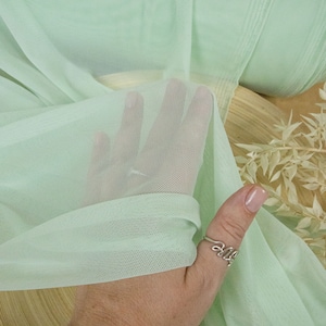 May include: Pale green sheer mesh fabric with a fine, even weave. The fabric is draped over a wooden surface and held in a hand wearing a silver ring.