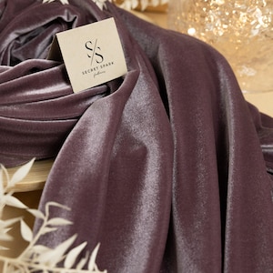 May include: A close-up of a deep purple velvet fabric with a subtle sheen. The fabric is folded and draped, showcasing its texture. A small, white logo with the text "SECRET SPARK fabrics" is visible on the fabric.