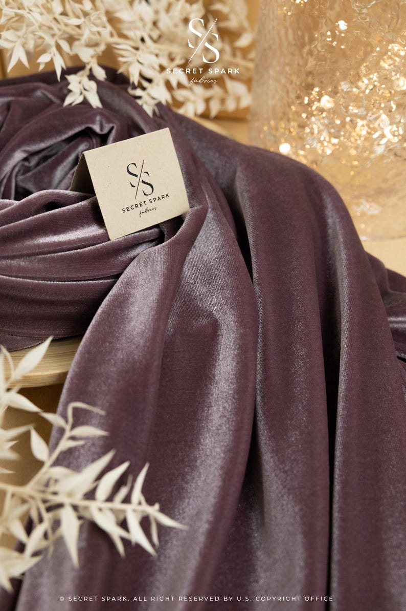 May include: A close-up of a deep purple velvet fabric with a subtle sheen. The fabric is folded and draped, showcasing its texture. A small, white logo with the text "SECRET SPARK fabrics" is visible on the fabric.