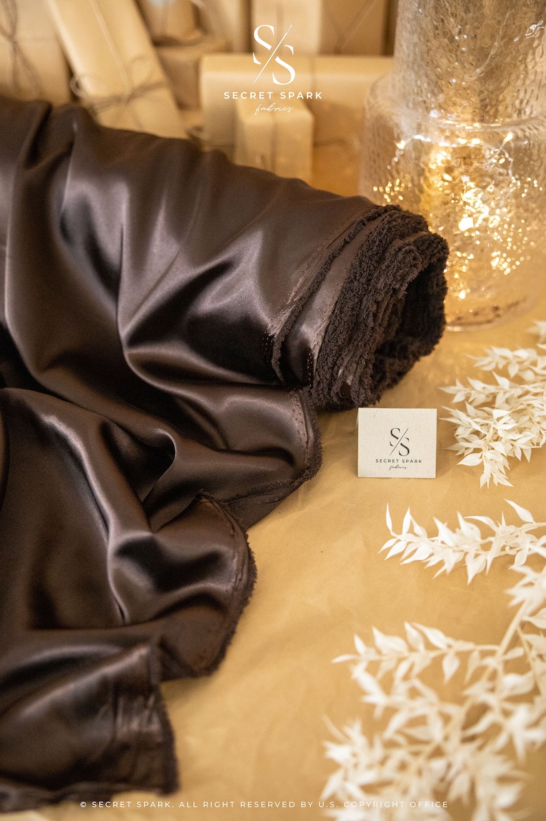 May include: A roll of dark brown satin fabric with a soft, silky texture. The fabric is folded and rolled up, with the edges of the fabric visible. The fabric is on a brown surface with white decorative branches.