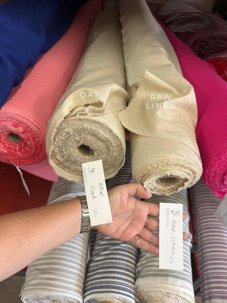 May include: Two rolls of fabric labeled OAT and RAW LINEN. The OAT roll is a light beige color, and the RAW LINEN roll is a slightly darker, natural tan shade. Both rolls have frayed edges and are positioned next to other colorful fabric rolls.