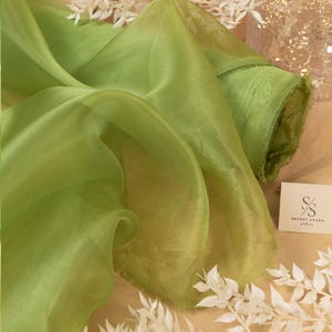 May include: A roll of light green sheer fabric with a frayed edge. The fabric is folded and draped over a white branch with green leaves. The text "SECRET SPARK" is visible on a tag.