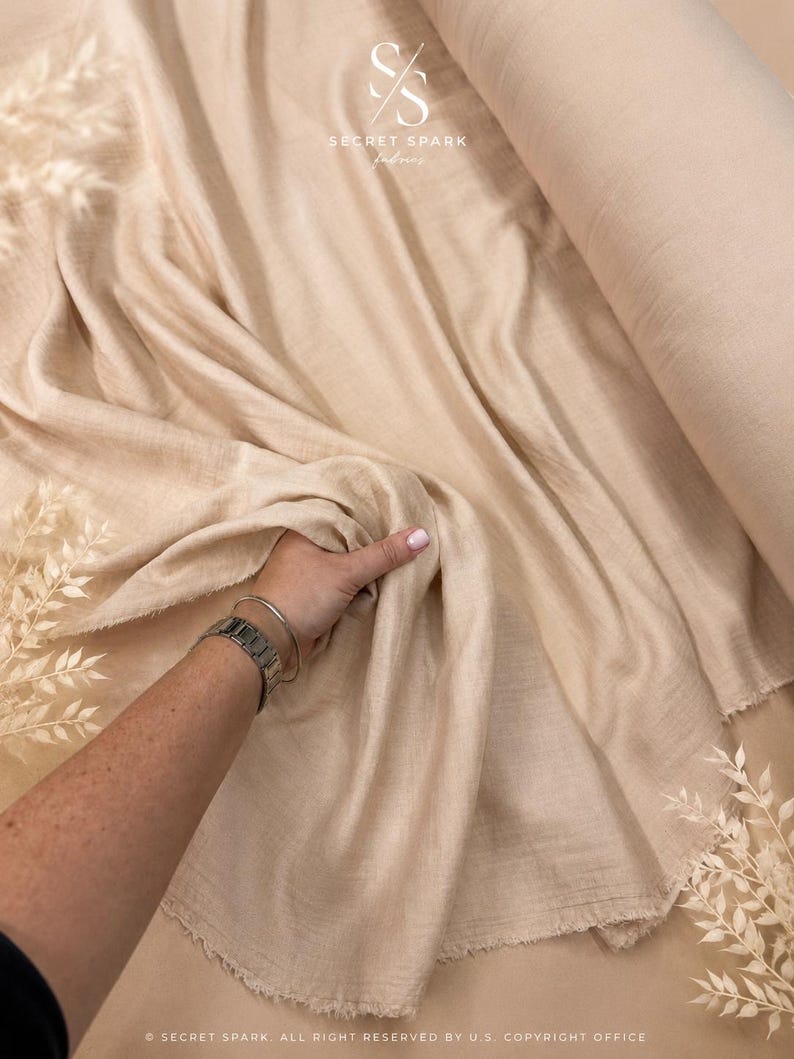 May include: A bolt of beige fabric with a soft, crinkled texture. The material is draped loosely, showing raw, frayed edges. A logo featuring the text SECRET SPARK fabrics is visible at the top, and copyright text appears at the bottom.