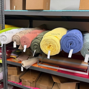 May include: A collection of fabric bolts arranged on a metal shelf, featuring colors labeled as lime, white, mystic beige, blush, dark olive, yellow, steel blue, and seafoam. Each roll is cylindrical with a visible texture and a small white tag attached.