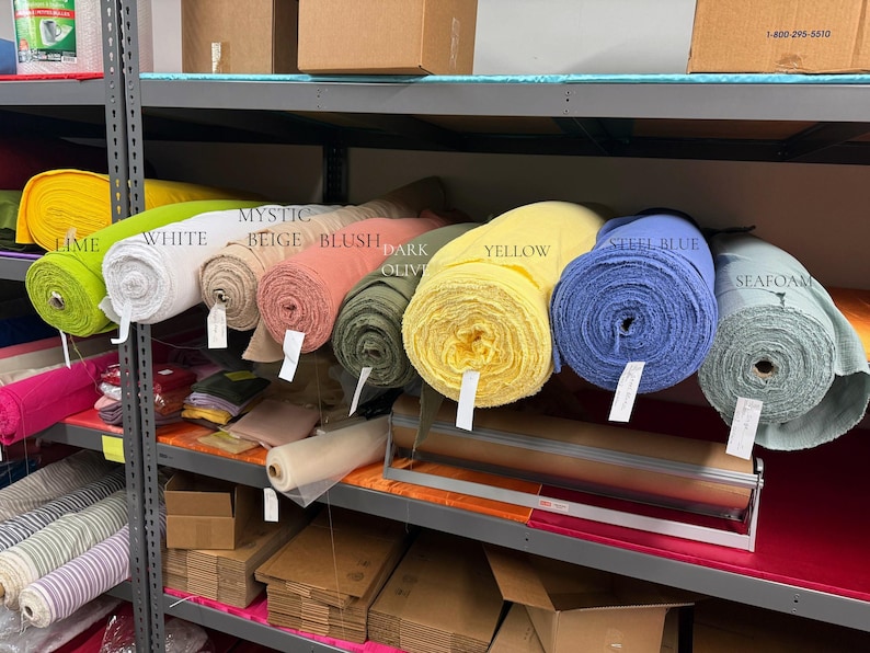 May include: A collection of fabric bolts arranged on a metal shelf, featuring colors labeled as lime, white, mystic beige, blush, dark olive, yellow, steel blue, and seafoam. Each roll is cylindrical with a visible texture and a small white tag attached.