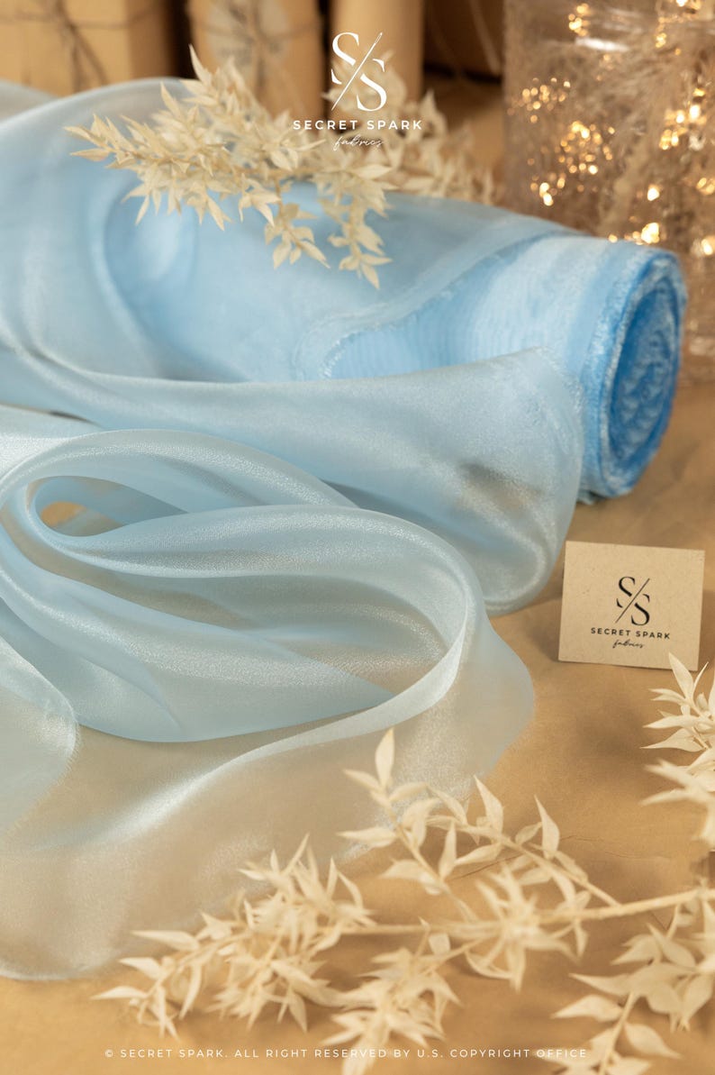 May include: A roll of light blue sheer fabric with a subtle shimmer. The fabric is rolled up and has a tag with the text "SECRET SPARK fabrics" on it.