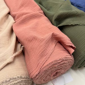 May include: Multiple rolls of crinkled muslin fabric in solid colors including beige, dusty rose, olive green, and blue. Each roll features a soft, textured surface with raw, frayed edges visible at the ends of the fabric bolts.
