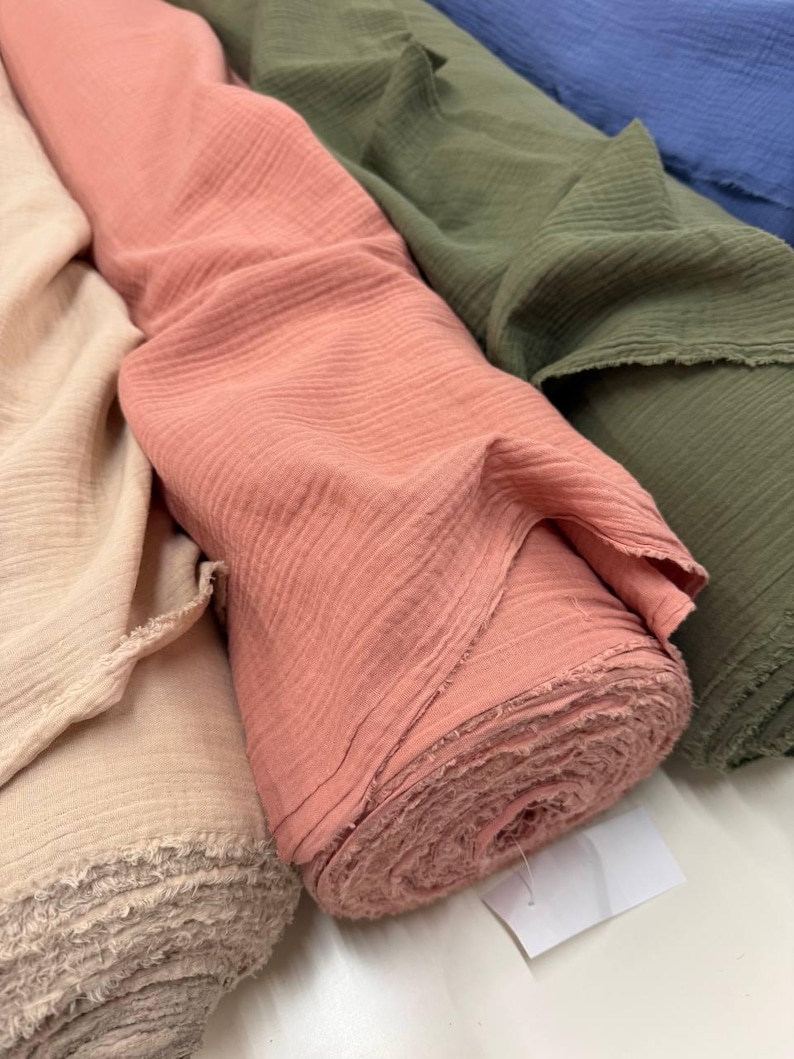 May include: Multiple rolls of crinkled muslin fabric in solid colors including beige, dusty rose, olive green, and blue. Each roll features a soft, textured surface with raw, frayed edges visible at the ends of the fabric bolts.