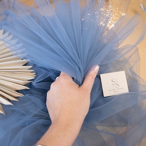 May include: A light blue tulle fabric with a hand holding the fabric. The fabric is folded in a fan shape. The fabric is labeled with the text "SECRET SPARK fabrics".