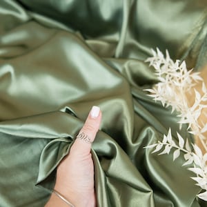 May include: A roll of smooth, sage green satin fabric. The fabric is draped over a surface and a hand is holding a corner of the fabric.