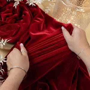 May include: A rich red velvet fabric is draped, with pleats and folds creating texture. The fabric is being held by hands, with a silver ring and bracelet visible. The background includes a decorative vase and white floral accents.