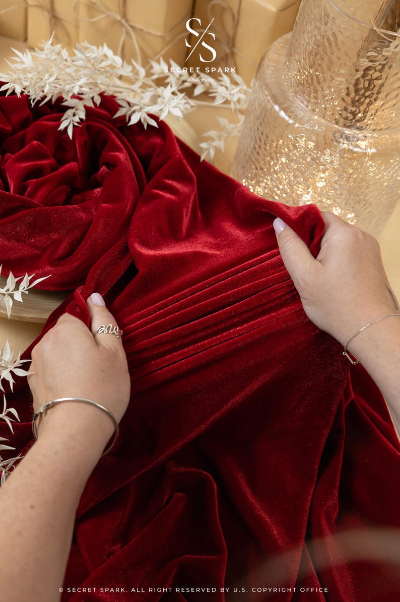 May include: A rich red velvet fabric is draped, with pleats and folds creating texture. The fabric is being held by hands, with a silver ring and bracelet visible. The background includes a decorative vase and white floral accents.